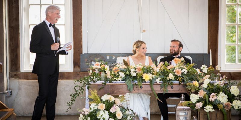 A newlywed couple sitting down while the bride's father gives a speech