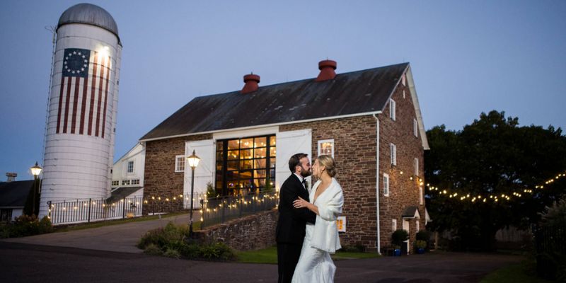 A newlywed couple standing in front of a barn and silo at night embracing and kissing