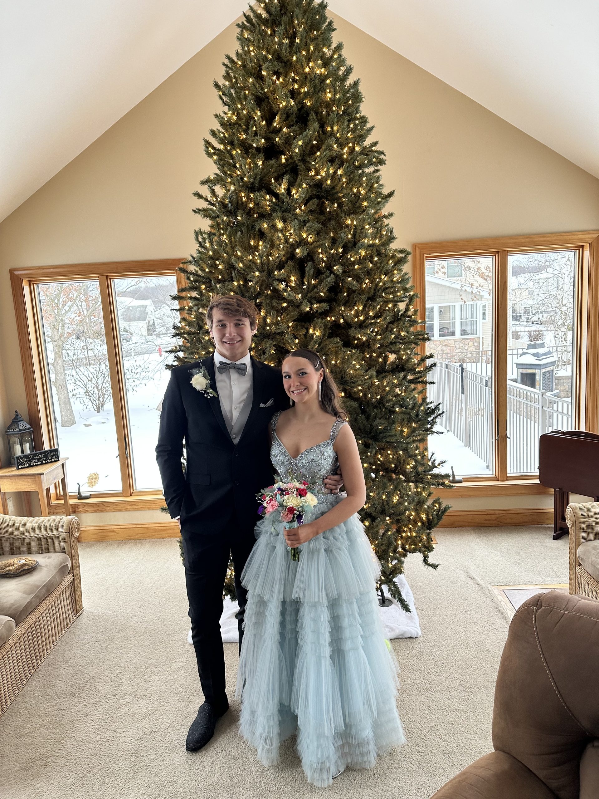 A young couple wearing formalwear standing smiling in front of a Christmas tree