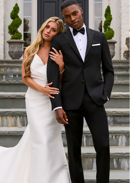 A bride in a white gown stands beside a groom in a black tuxedo on outdoor stone steps, with her arm wrapped around his, both looking at the camera with a serene expression.