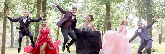 Six people dressed in formal prom attire are jumping and smiling outdoors among trees, celebrating and posing energetically on a sunny day.