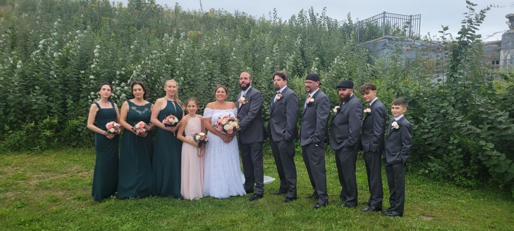 A wedding party stands outside on grass in front of tall greenery. The bride in a white dress is surrounded by bridesmaids in dark and blush dresses and groomsmen in gray suits with boutonnieres, all holding bouquets and smiling.