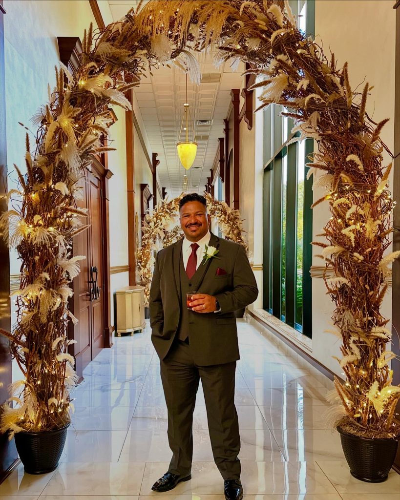A man in a dark suit stands smiling with a drink in hand under a decorative arch of dried grasses and flowers, inside a brightly lit hallway with shiny floors and large windows.