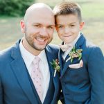 A smiling man and young boy, both wearing blue suits with boutonnieres, pose closely together outdoors. The boy has his arm around the mans shoulder, and they both look happy.