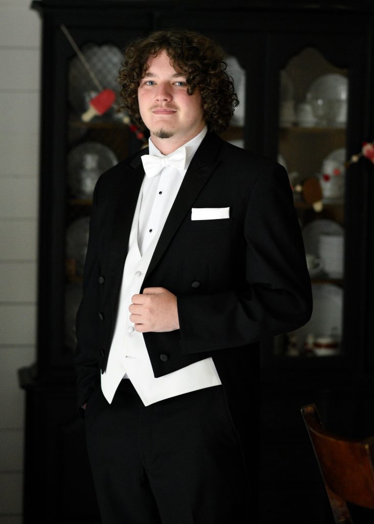 A young man with curly hair wears a formal black tuxedo with a white bow tie and vest, standing indoors in front of a dark china cabinet filled with dishes. He looks at the camera with a slight smile.