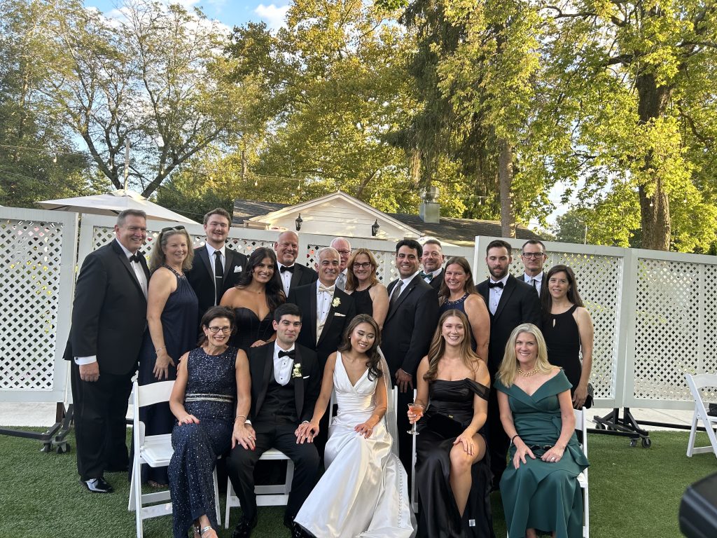 A group of people dressed in formal attire, including tuxedos and gowns, pose together outdoors in front of a white fence and trees. Some are seated while others stand behind them, smiling at the camera.