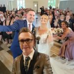 A bride and groom smile with their wedding party for a group selfie at their indoor ceremony, with guests seated behind them and everyone looking joyful.