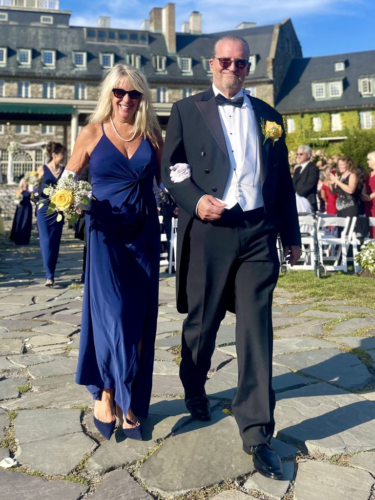 A smiling woman in a navy blue dress and sunglasses walks arm-in-arm with a man in a tuxedo with a yellow boutonniere at an outdoor wedding, with guests and a stone building in the background.