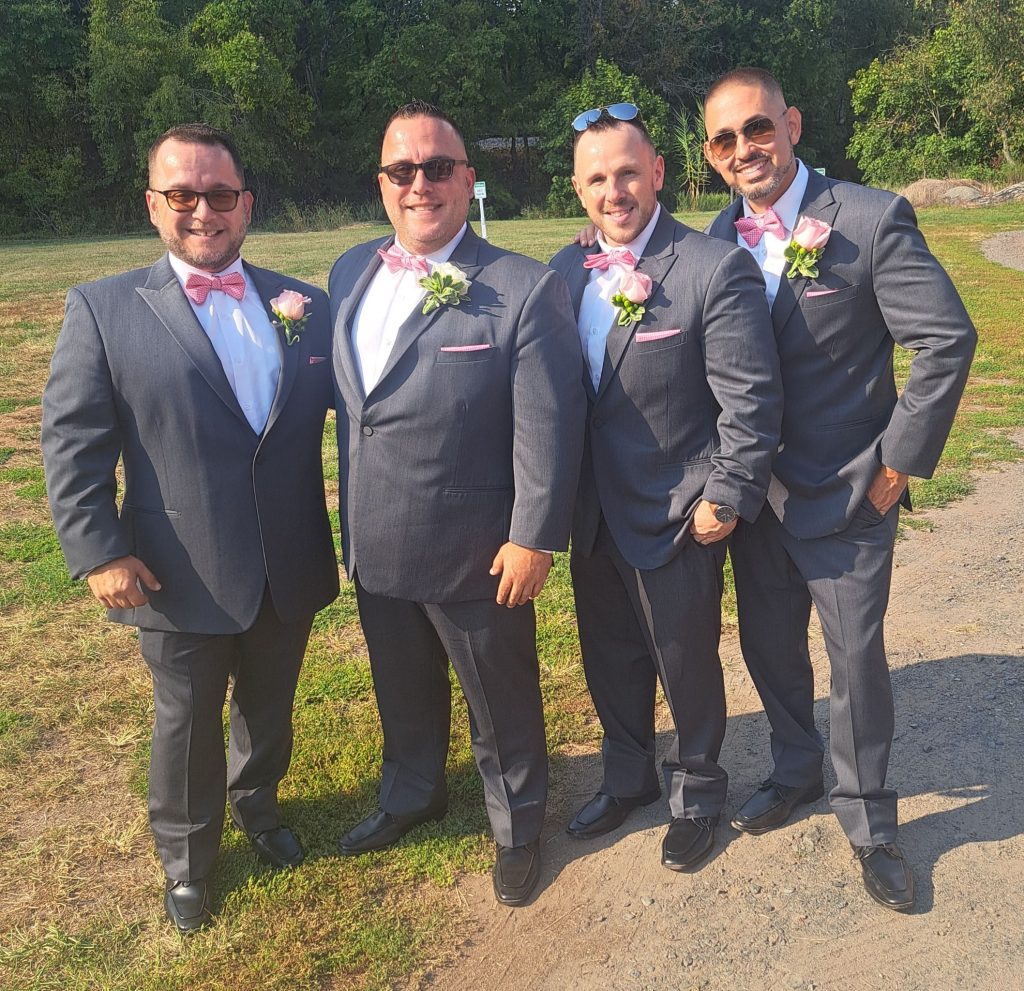Four men in matching gray suits, pink bow ties, and pink boutonnieres stand together outdoors on a sunny day, smiling. Trees and grass are visible in the background.