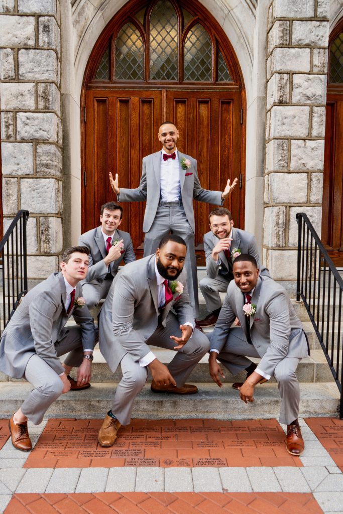 Six men in light gray suits pose playfully on church steps in front of wooden doors, with the groom standing in the center back while the others crouch or kneel, all smiling and laughing together.