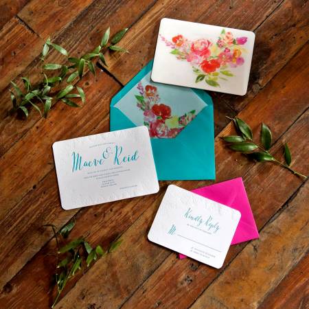 Wedding invitations and RSVP cards with teal and pink envelopes are arranged on a wooden floor with green leaves. One envelope is open, showing a floral lining; another has a floral design on the outside.