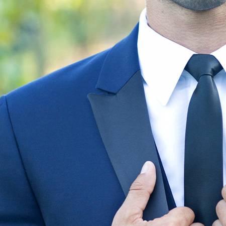 A person wearing a dark blue suit, white dress shirt, and black tie adjusts their tie with one hand. The image is cropped to show only the upper chest and part of the face, outdoors with a blurred background.
