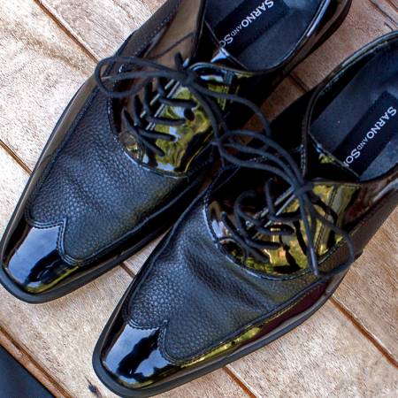 A pair of shiny black dress shoes with textured and glossy sections, featuring black laces, placed on a wooden surface.