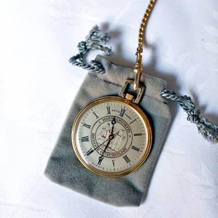A gold pocket watch with a roman numeral face and chain rests on a small gray velvet pouch, placed on a white surface.