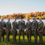 Seven men in matching brown suits, one wearing a cowboy hat, stand together on grass by a lake with colorful autumn trees in the background, posing and smiling for a group photo.