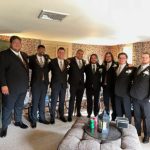 A group of eight men in black suits with boutonnieres stand arm-in-arm in a living room with floral wallpaper, smiling at the camera. There are drinks and decorative items on the ottoman in front of them.