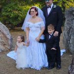 A bride and groom pose outdoors with two young children, all dressed formally. The bride wears a white gown and veil, the groom a black suit with a top hat and sunglasses. Trees and large rocks are in the background.