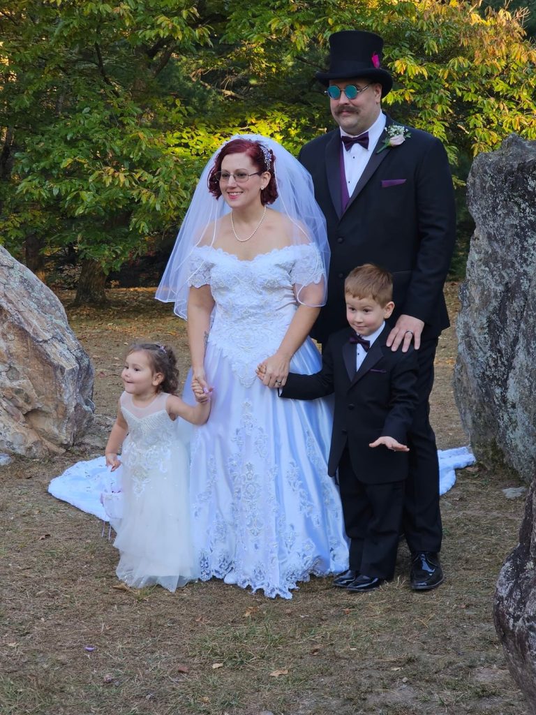 A bride and groom pose outdoors with two young children, all dressed formally. The bride wears a white gown and veil, the groom a black suit with a top hat and sunglasses. Trees and large rocks are in the background.