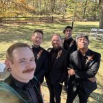 Five men dressed in black suits and boutonnières stand outdoors on grass, smiling for a selfie. Behind them are trees with autumn leaves, a wooden fence, and white chairs set up for an event.