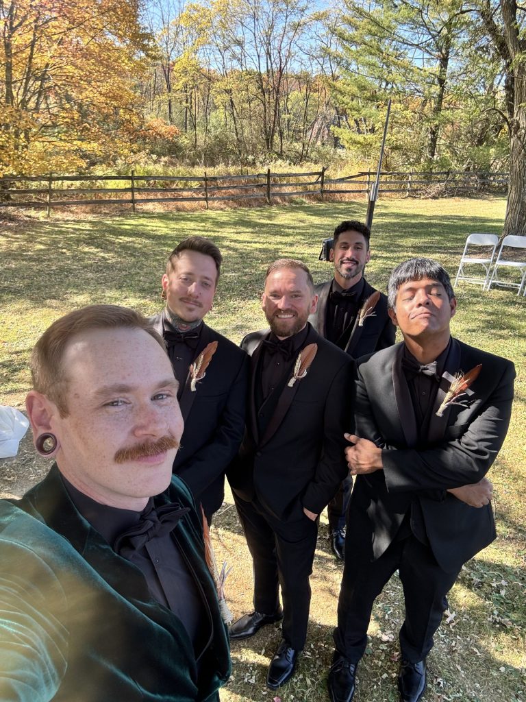 Five men dressed in black suits and boutonnières stand outdoors on grass, smiling for a selfie. Behind them are trees with autumn leaves, a wooden fence, and white chairs set up for an event.