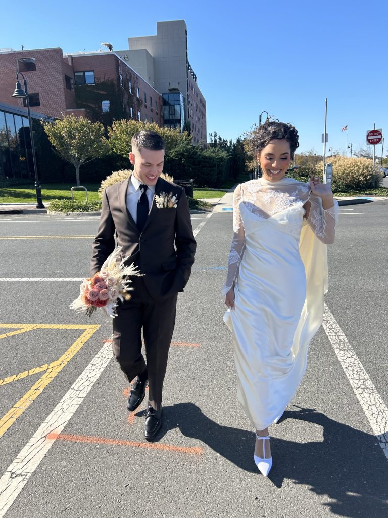 A smiling bride in a long white dress and a groom in a dark suit walk together across a sunlit street, holding hands with a bouquet and enjoying their wedding day under a clear blue sky.