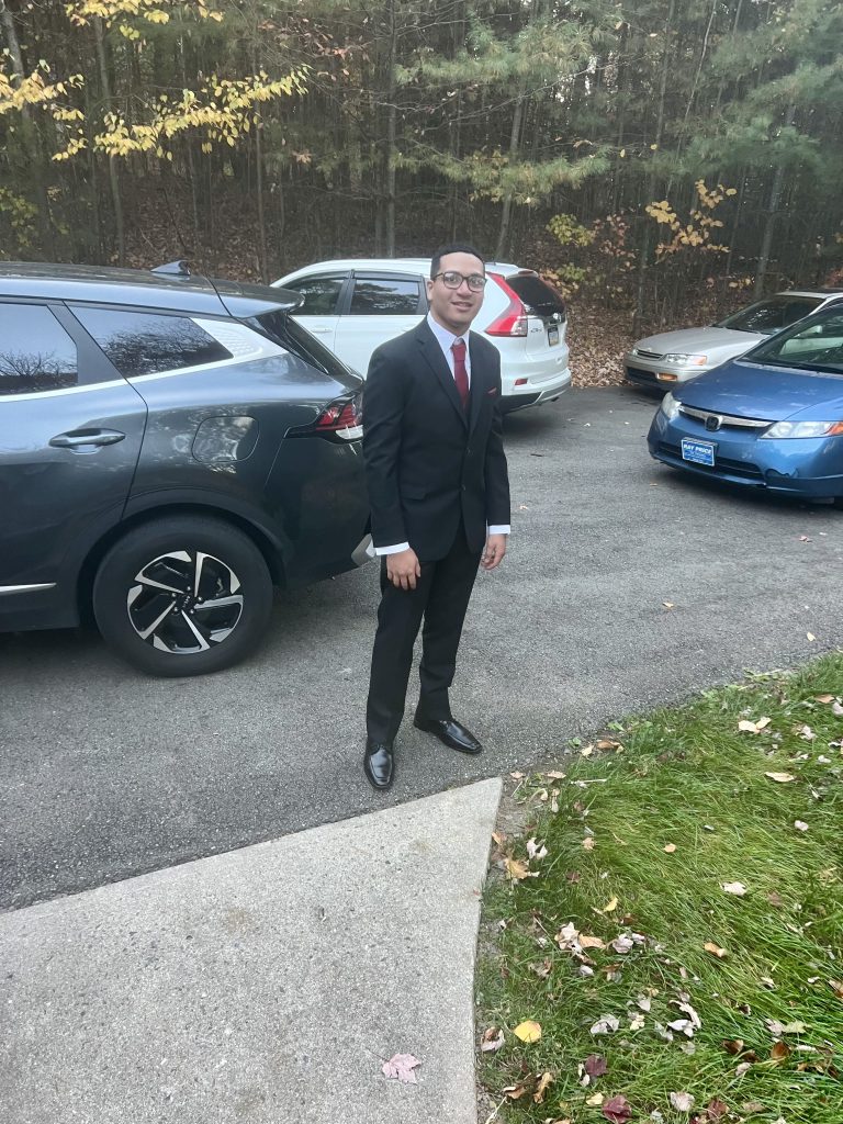 A man in a black suit, white shirt, and red tie stands on a driveway next to parked cars. Autumn leaves are scattered on the ground, and trees with fall foliage are in the background.