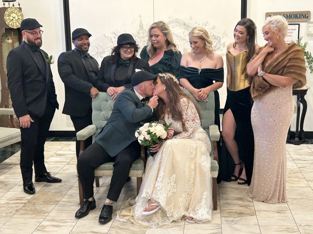 A bride and groom kiss while sitting, surrounded by six smiling friends dressed in formal and semi-formal attire. The bride holds a bouquet, and everyone poses cheerfully in an elegant indoor setting.