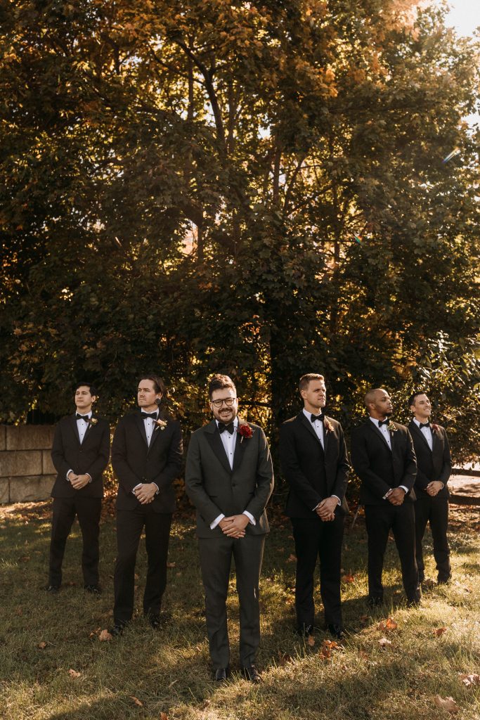 Six men in tuxedos stand in a row outdoors on grass, with trees and sunlight in the background. The man in the center is slightly ahead of the others and has a red rose on his lapel.