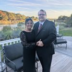 A couple dressed formally in black attire stands together on a wooden deck by a lake, smiling at the camera. Trees with fall foliage and a partly cloudy sky are visible in the background.