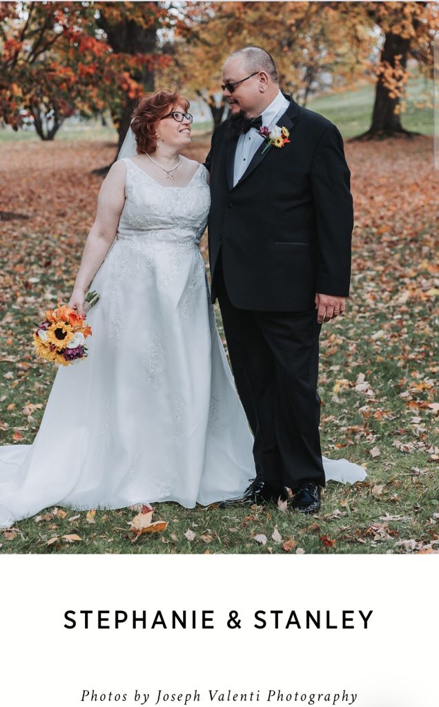 A bride in a white dress and a groom in a black tuxedo smile at each other outdoors in a park with autumn leaves. She holds a bouquet of sunflowers and other fall flowers. Text below reads STEPHANIE & STANLEY.