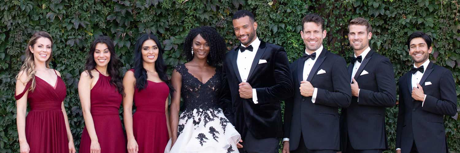 A wedding party poses outdoors in front of leafy greenery. The bride wears a black and white gown, bridesmaids are in burgundy dresses, and the groom and groomsmen wear black tuxedos with bow ties. Everyone is smiling.