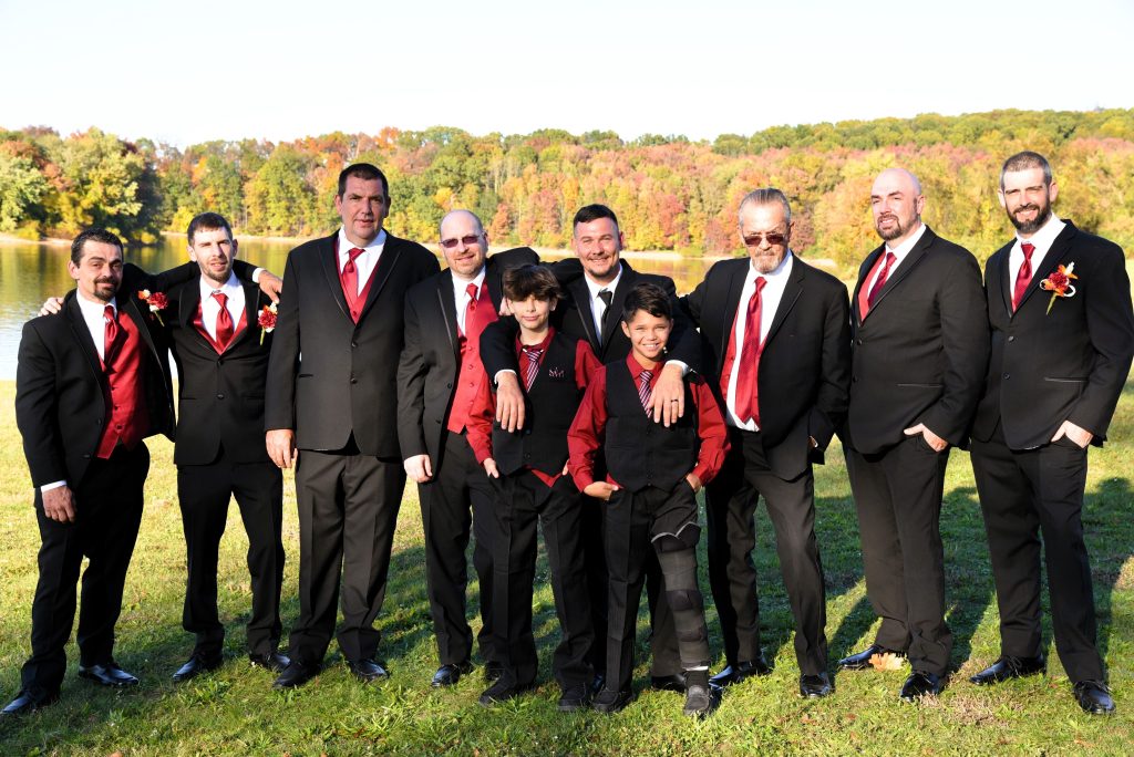 A group of ten men and boys dressed in black suits, some with red vests and ties, stand smiling together outdoors on grass with trees and a lake in the background on a sunny day.
