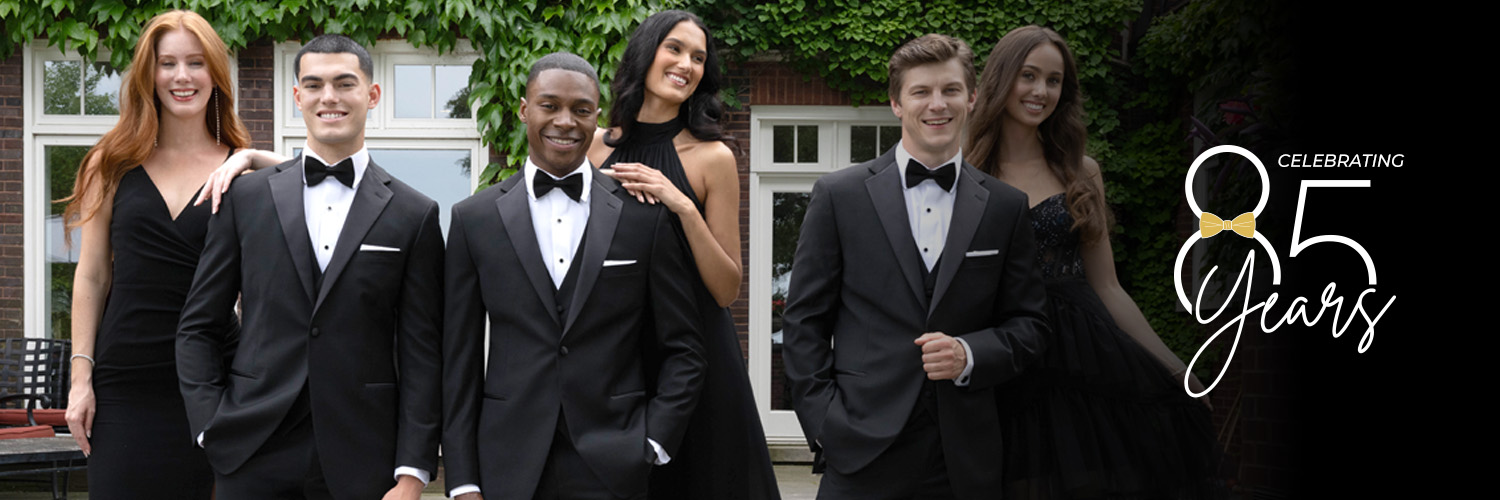 Five young adults in formal black attire, three men in tuxedos and two women in black dresses, smile outdoors in front of ivy-covered windows. Text reads, “Celebrating 85 Years.”.