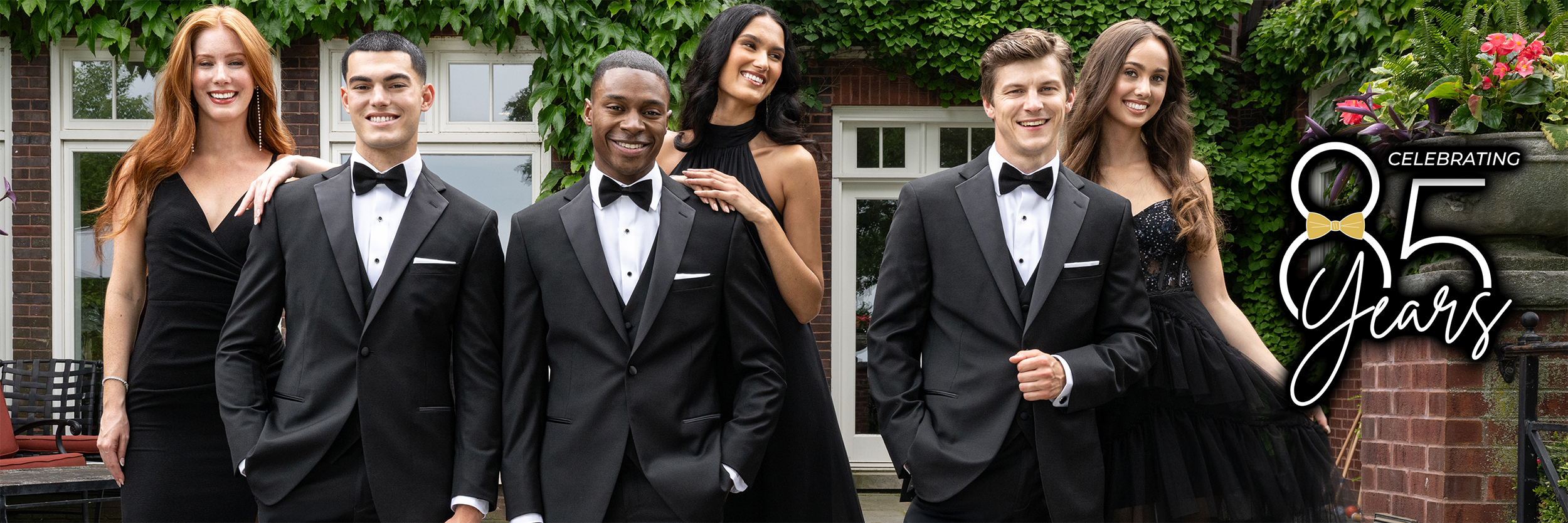 Five young adults in formal black attire, three men in tuxedos and two women in black dresses, smile outdoors in front of ivy-covered windows. Text reads, “Celebrating 85 Years.”