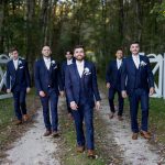 Six men in navy blue suits and brown shoes walk confidently down a tree-lined path with white gates in the background, smiling and dressed formally, likely for a wedding.