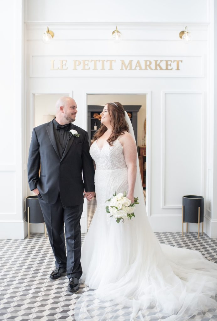 A bride and groom stand holding hands, smiling at each other. The bride wears a white gown and holds a bouquet; the groom wears a black suit. They are indoors in front of a sign that reads LE PETIT MARKET.