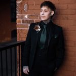 A person with short hair and tattoos, wearing a black suit with a dark green shirt and tie, stands against a brick wall, gazing to the side with hands in their pockets.