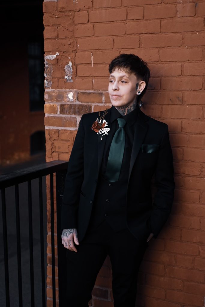 A person with short hair and tattoos, wearing a black suit with a dark green shirt and tie, stands against a brick wall, gazing to the side with hands in their pockets.
