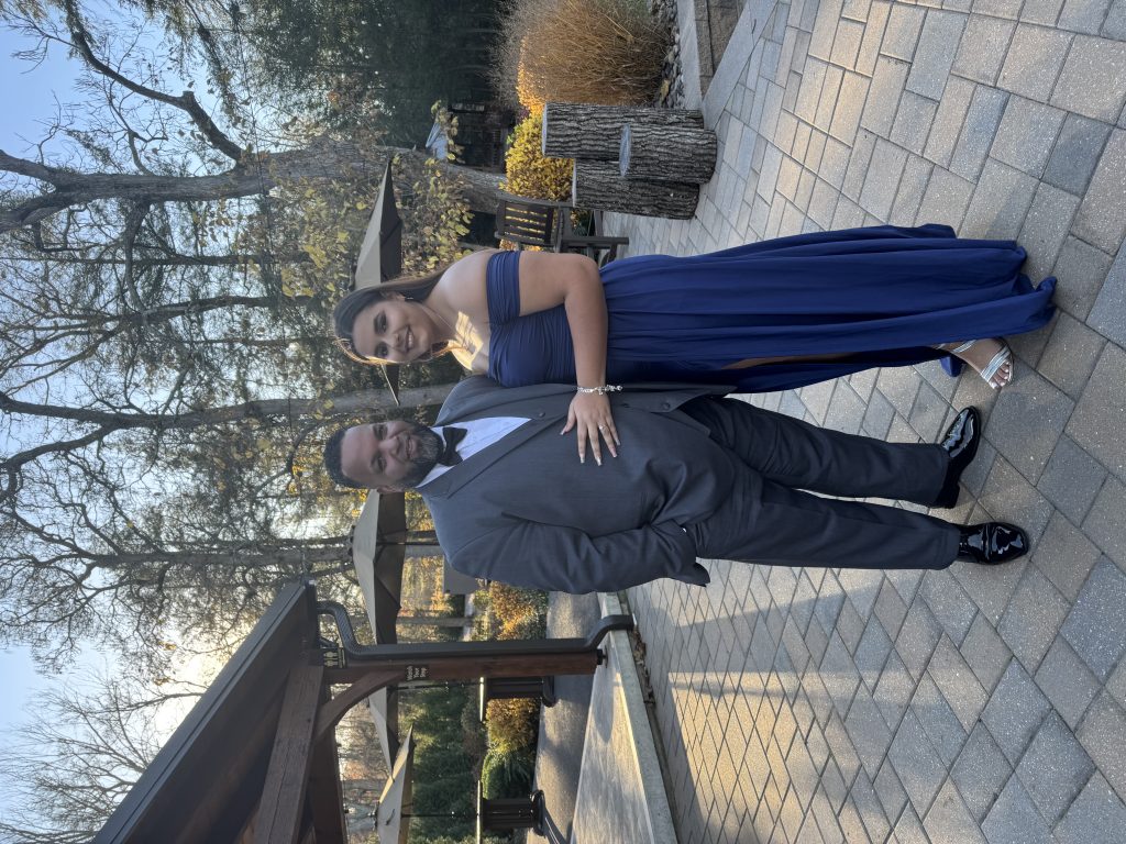A woman in a blue off-shoulder gown stands next to a man in a gray suit and bow tie, both smiling outdoors on a stone patio with trees and wooden structures in the background.