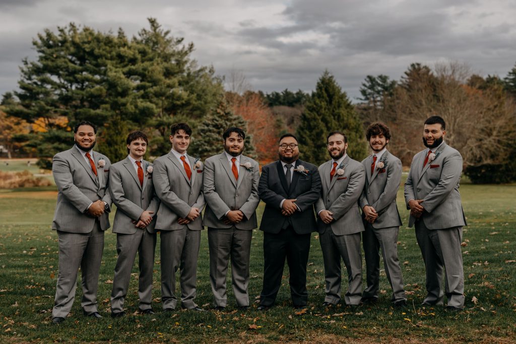 Eight men stand in a row outdoors on grass, seven in light gray suits with orange ties and boutonnieres, and one in a dark suit, with trees and cloudy sky in the background.