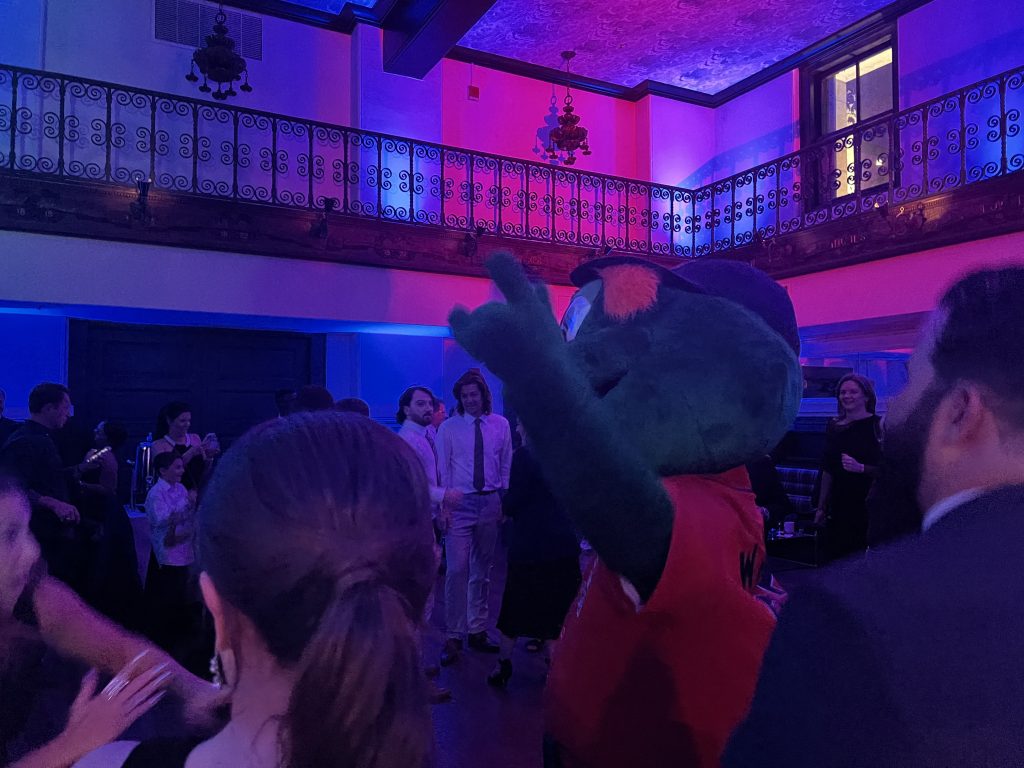 A crowd of people in formal attire dance in a dimly lit room with blue and purple lights. A person in a large green mascot costume stands in the center, raising an arm. Ornate railings and high ceilings are visible in the background.