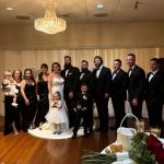 A bride and groom stand together with children and a wedding party dressed in formal black attire, posing for a group photo under a chandelier in a warmly lit reception hall.