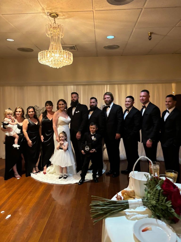 A bride and groom stand together with children and a wedding party dressed in formal black attire, posing for a group photo under a chandelier in a warmly lit reception hall.