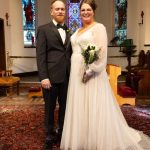 A bride in a white gown holding a bouquet stands next to a groom in a black suit. They pose and smile in a church with stained glass windows and ornate decor, standing on a patterned red carpet.