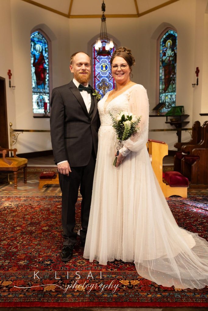 A bride in a white gown holding a bouquet stands next to a groom in a black suit. They pose and smile in a church with stained glass windows and ornate decor, standing on a patterned red carpet.