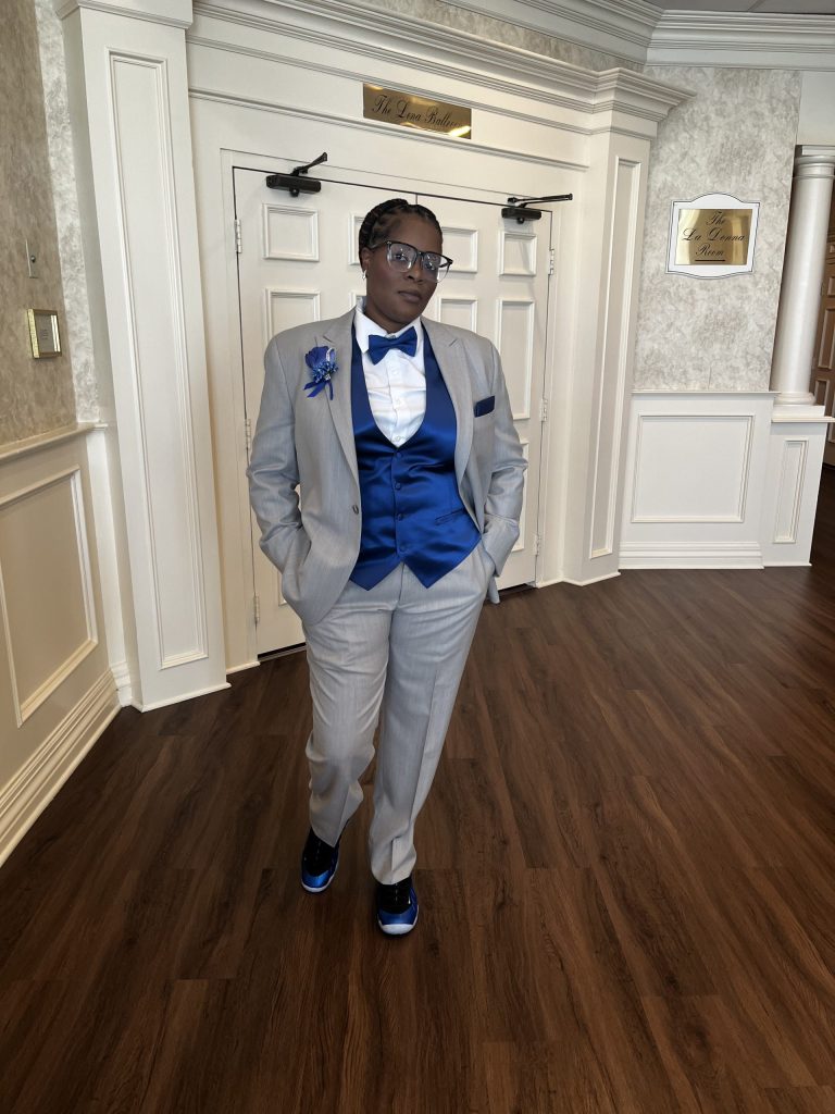 A person in a light gray suit with a royal blue vest, bow tie, and shoes stands confidently indoors. Their hands are in their pockets, and they are wearing glasses. The background features white paneled walls and double doors.