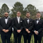 Four men dressed in black tuxedos with bow ties stand side by side outdoors in front of green trees, smiling at the camera on a cloudy day.