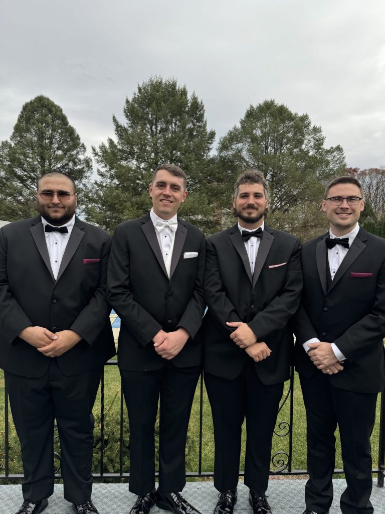 Four men dressed in black tuxedos with bow ties stand side by side outdoors in front of green trees, smiling at the camera on a cloudy day.