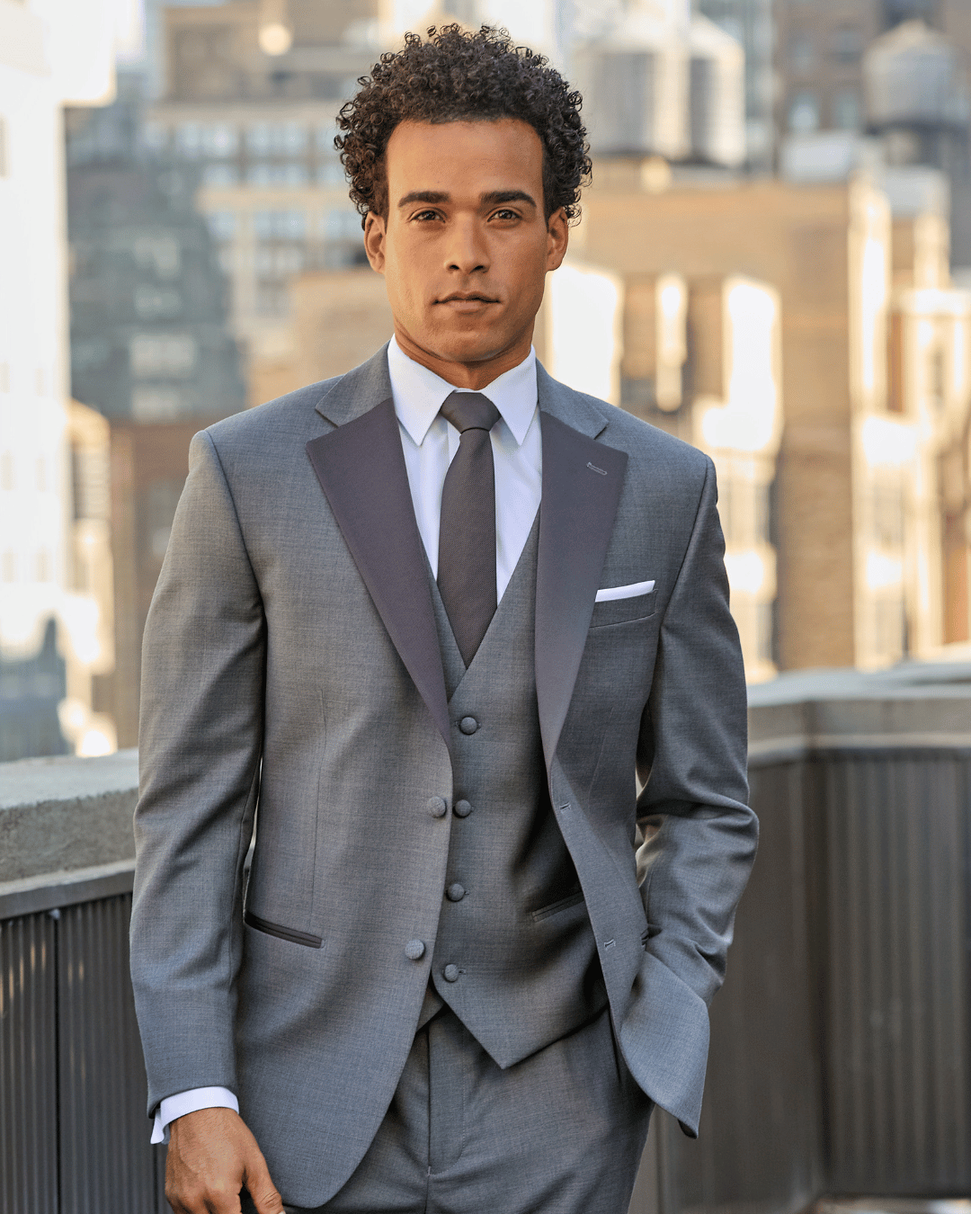 A man in a gray suit, white dress shirt, black bow tie, and sunglasses stands confidently on a city rooftop with blurred buildings in the background.