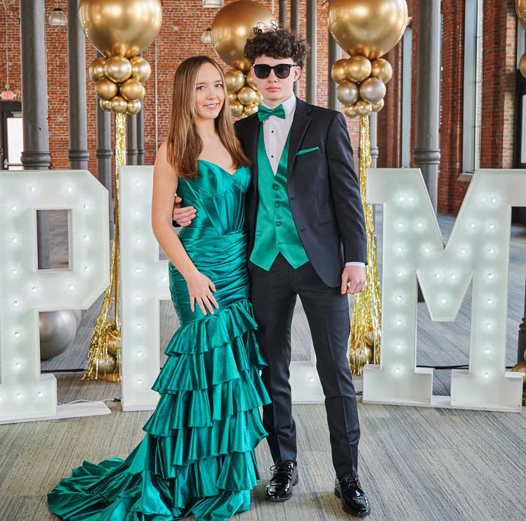 A young couple poses in front of large illuminated “PROM” letters and gold balloon decorations. The girl wears a green ruffled gown, and the boy wears a black suit with a matching green vest and bow tie.